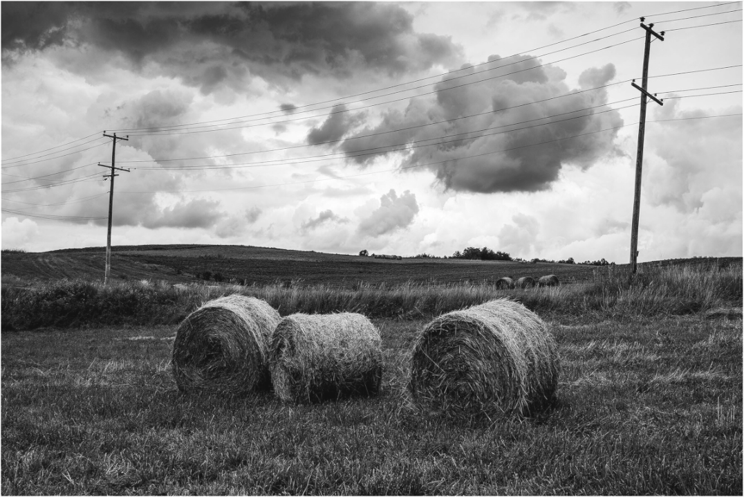 Black and white photograph of three hay bales in an open field beneath a dramatic sky—minimalist rural image evoking balance, weight, and quiet space.