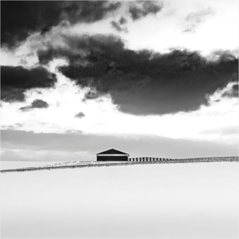 Black and white photograph of a solitary barn beneath dramatic sky—minimalist rural composition conveying quiet, distance, and space.