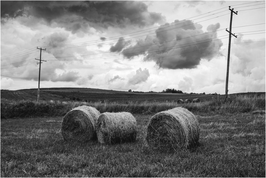 Black and white photograph of three hay bales in an open field beneath a dramatic sky—minimalist rural image evoking balance, weight, and quiet space.

